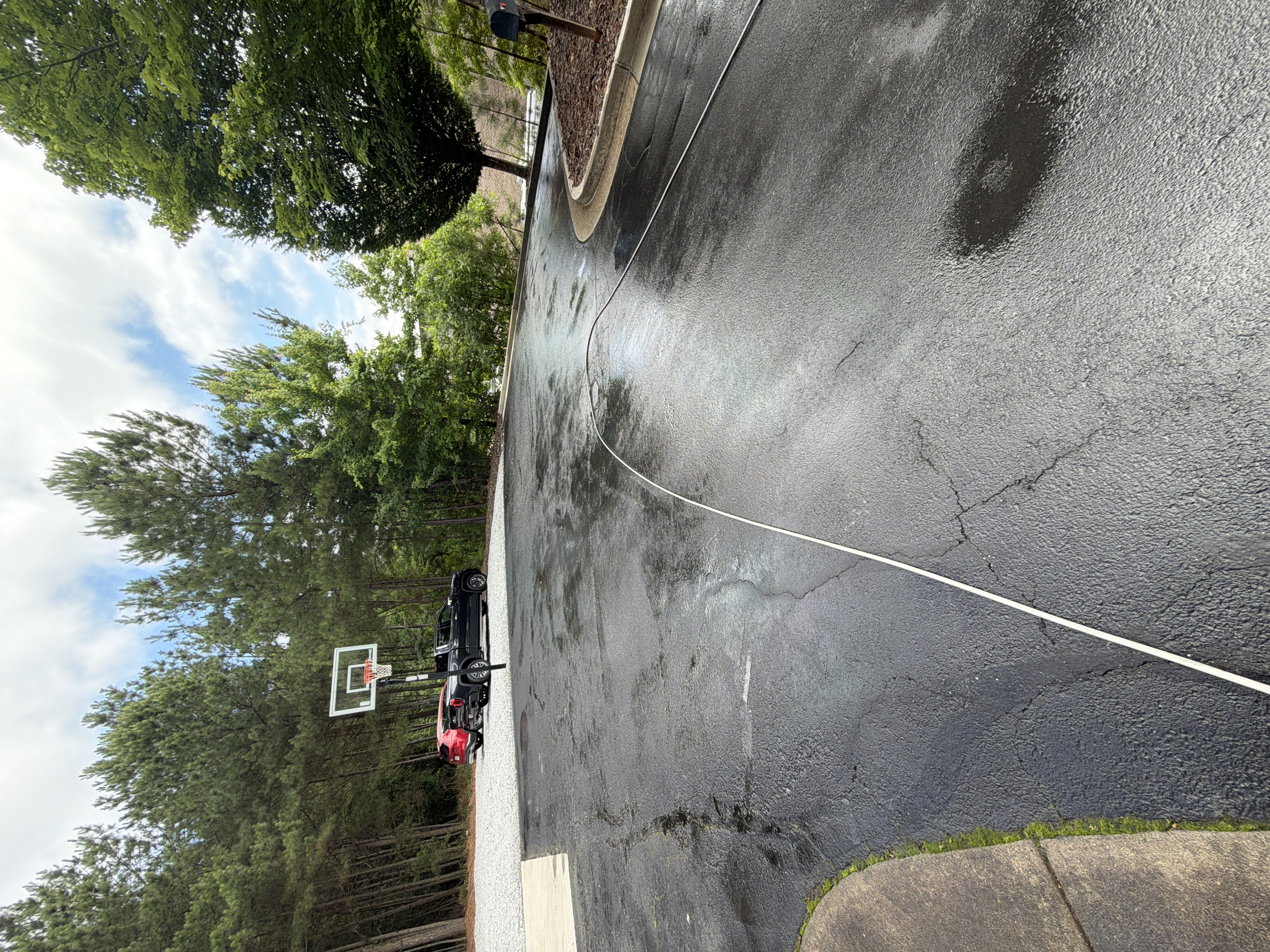 Wet asphalt driveway and basketball court area during pressure washing in Alpharetta, GA, with hose visible and trucks parked in the background