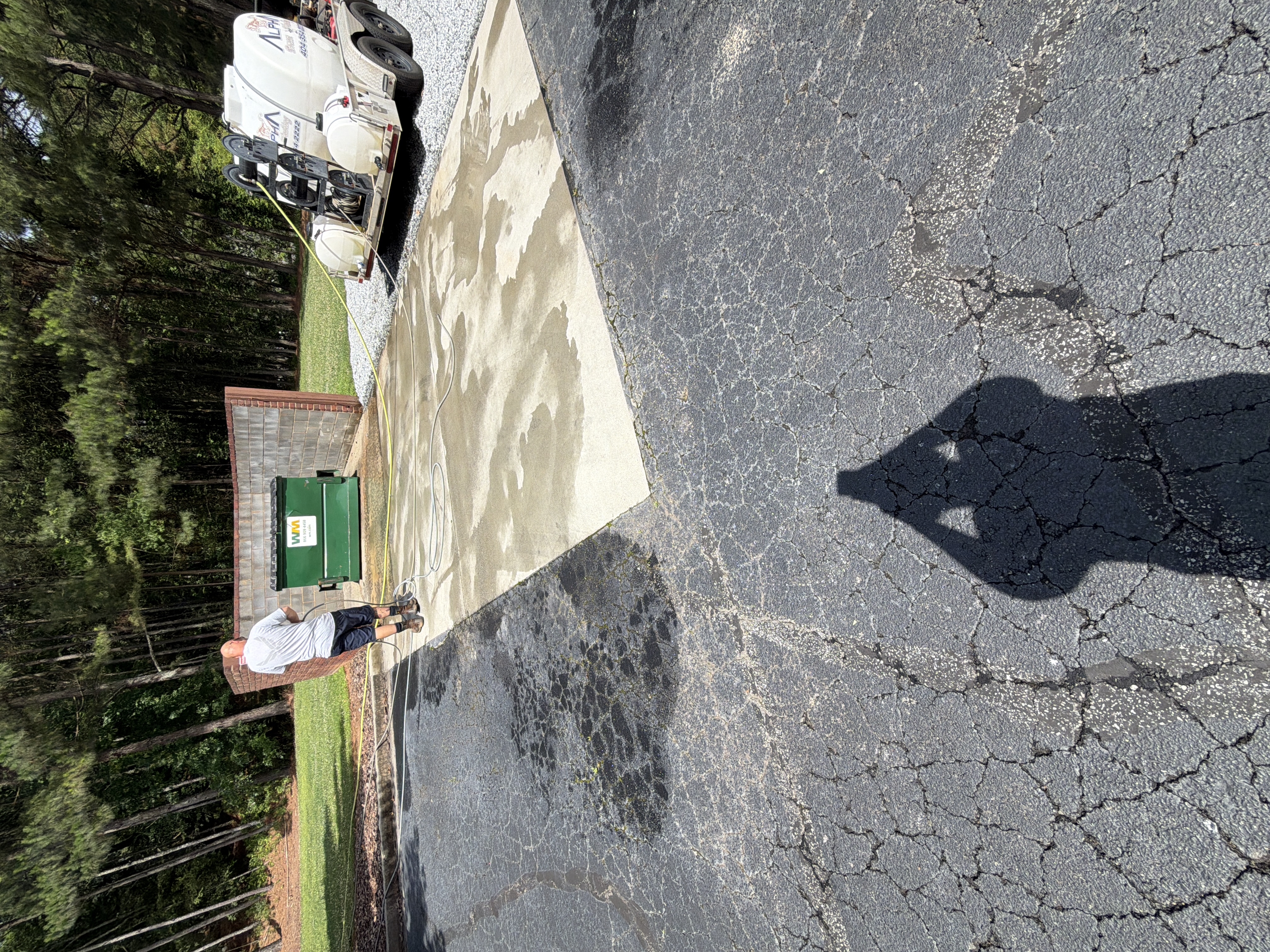 Alpha Pressure Washing technician cleaning a concrete pad near a dumpster enclosure in Alpharetta, GA, with the service trailer visible beside the building