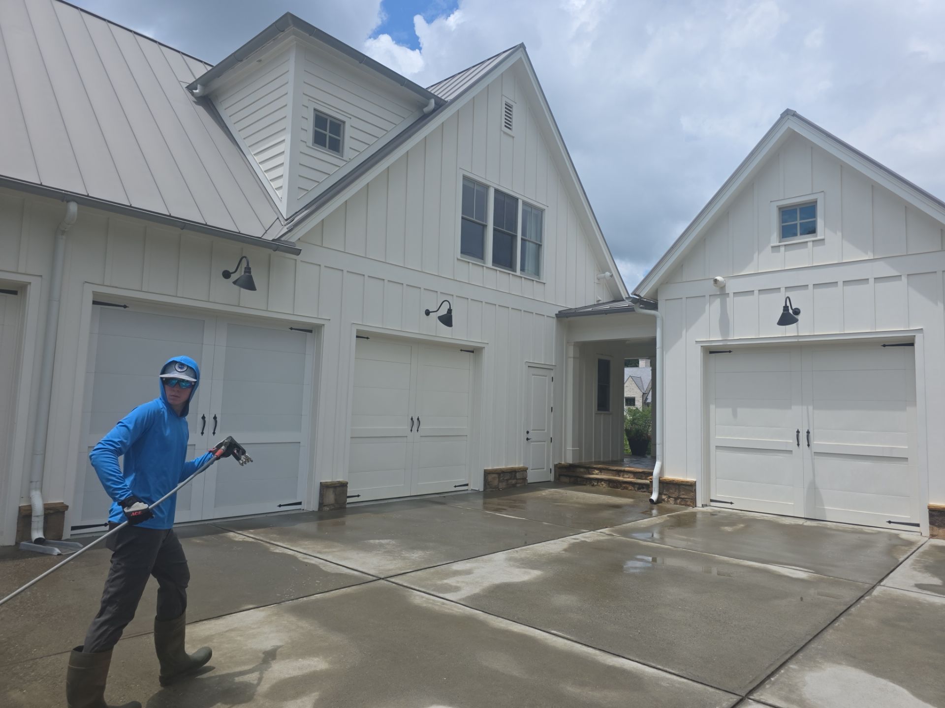 Technician pressure washing a concrete driveway in front of a white board-and-batten home with a three-car garage in Milton, GA.