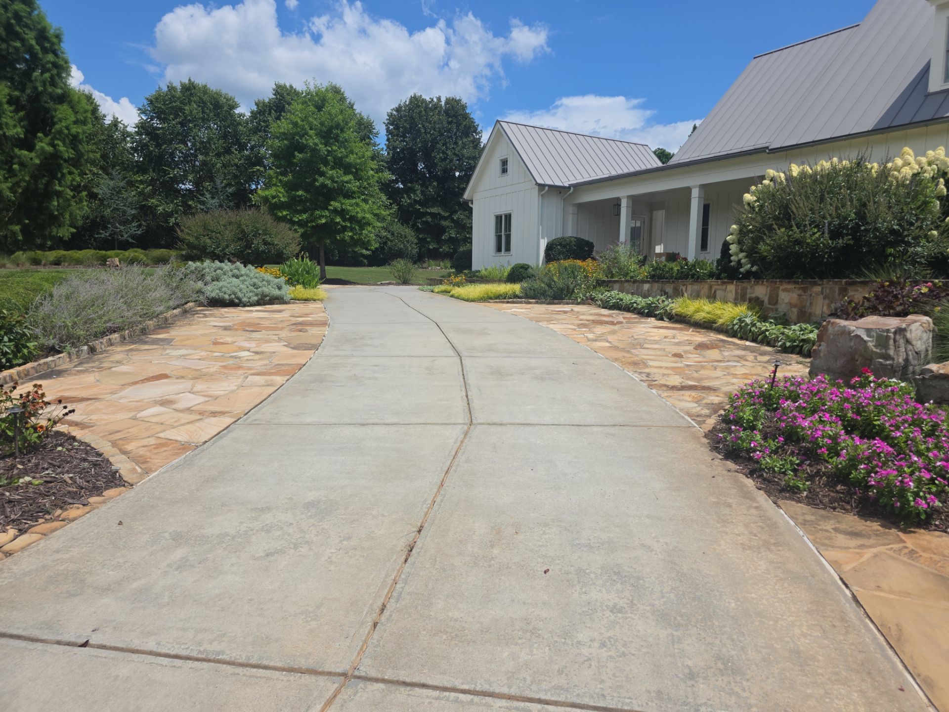 Curved concrete driveway leading to a white farmhouse flanked by flagstone shoulders and garden beds in Milton, GA.