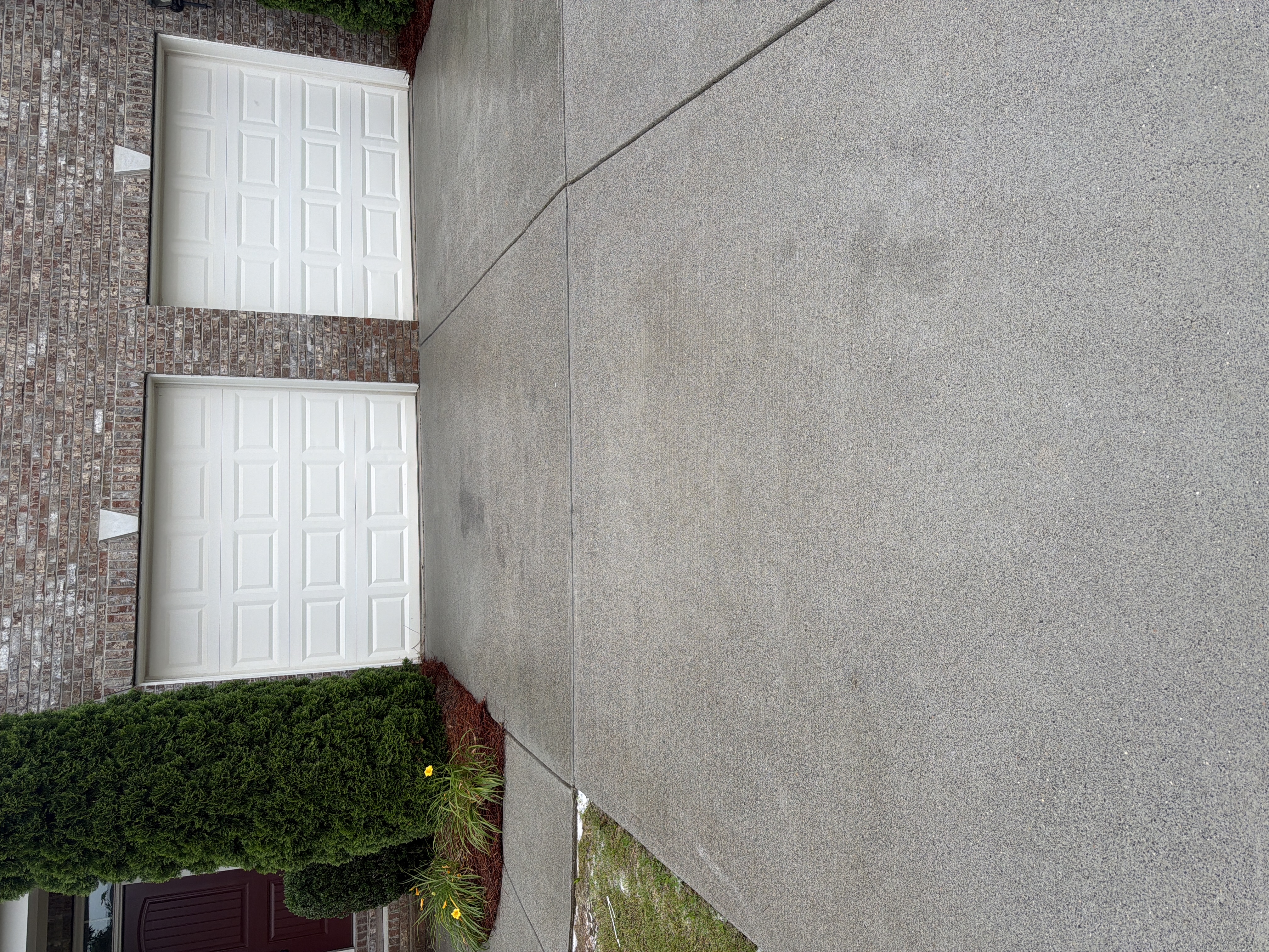 Pressure-washed two-car concrete driveway in Cumming, GA with brick home and cedar bushes, surface looking clean and uniform